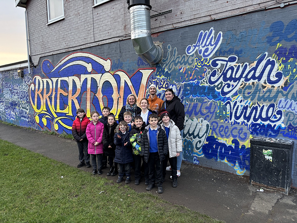 A group of children and adults stand in front of a colourful wall mural that reads “Brierton,” with stylised names painted across the background.
