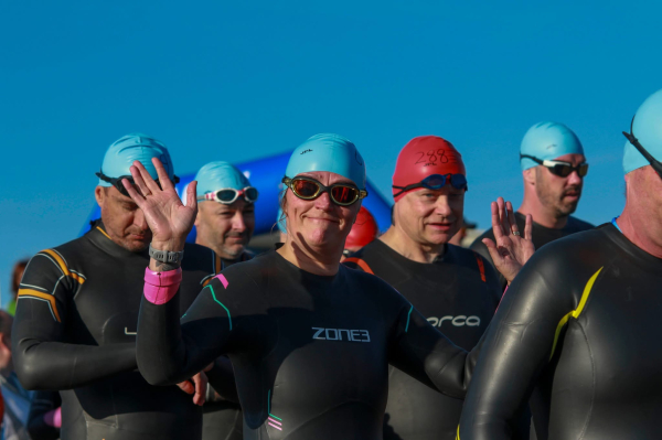 Swimmers in wetsuits and colorful caps, some with hands raised, preparing for a race under a clear blue sky.