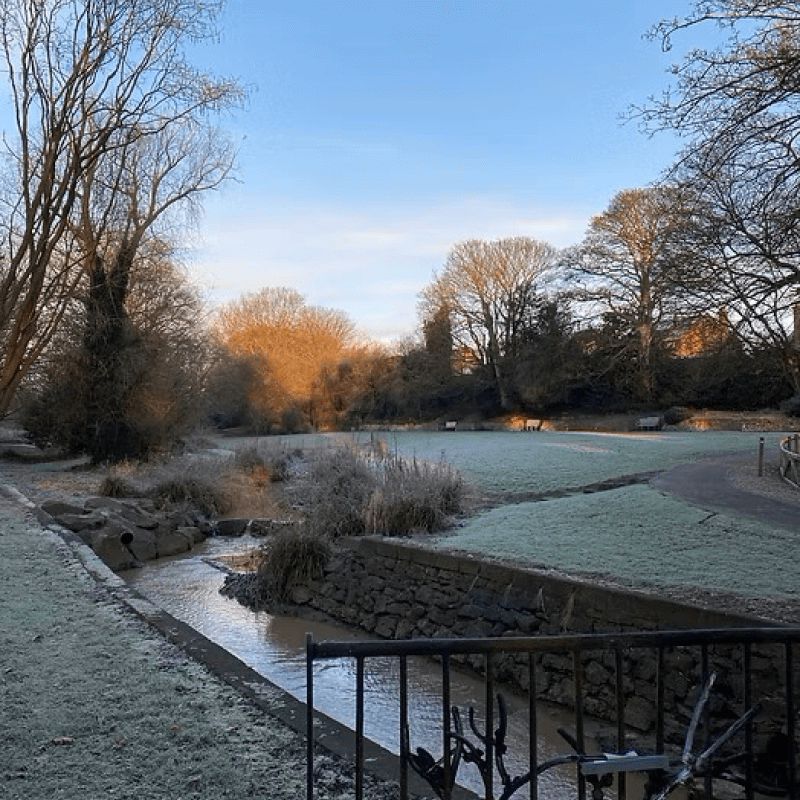 A frosty winter park with a small stream flowing under a stone bridge, leafless trees, benches along a path, and sunlight casting an orange glow on distant trees.