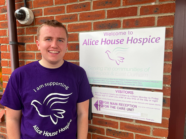 A person in a purple “I am supporting Alice House Hospice” T‑shirt stands beside Alice House Hospice signage on a brick wall.