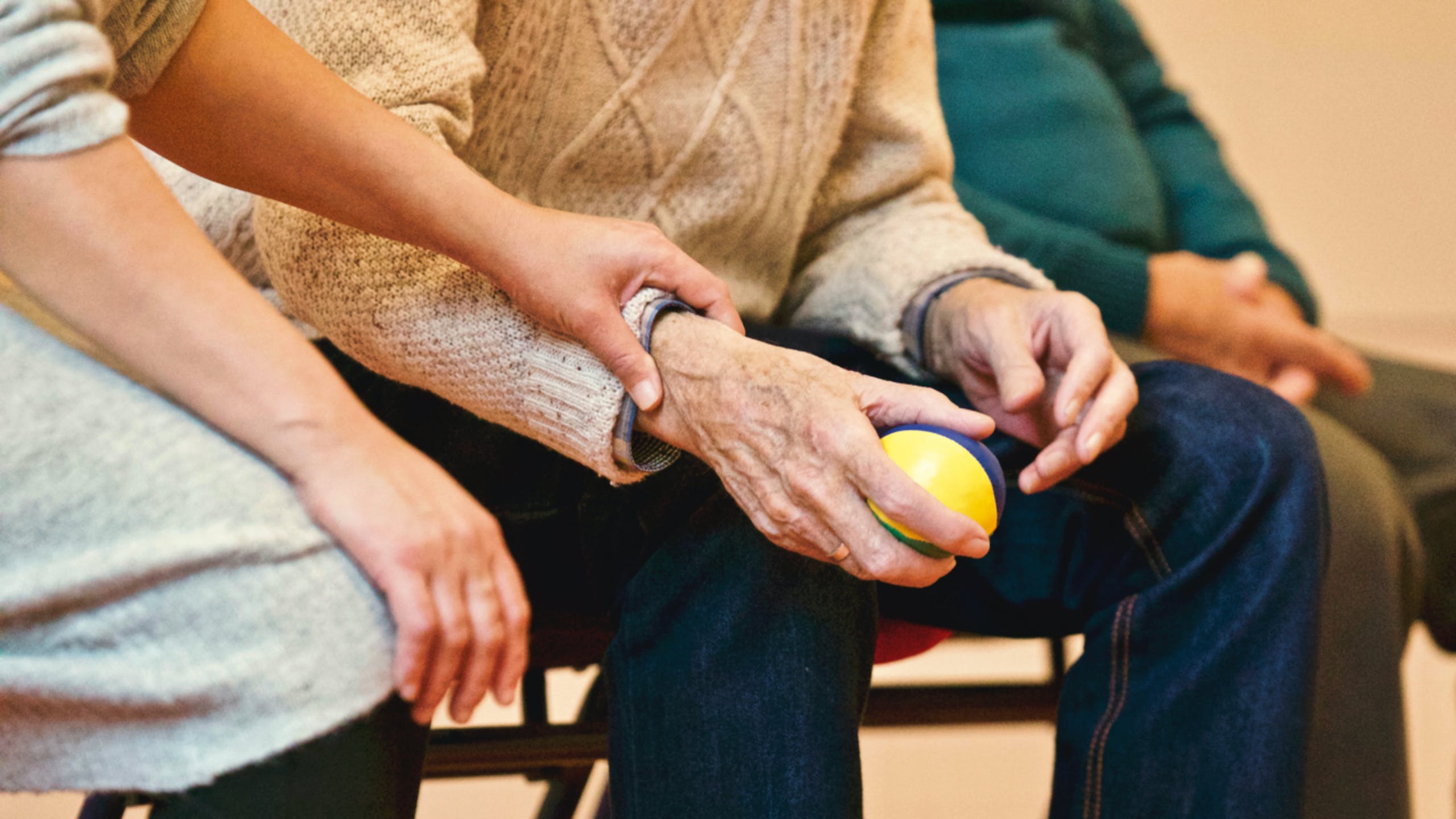 Elderly person holding a small ball, assisted by another individual indoors
